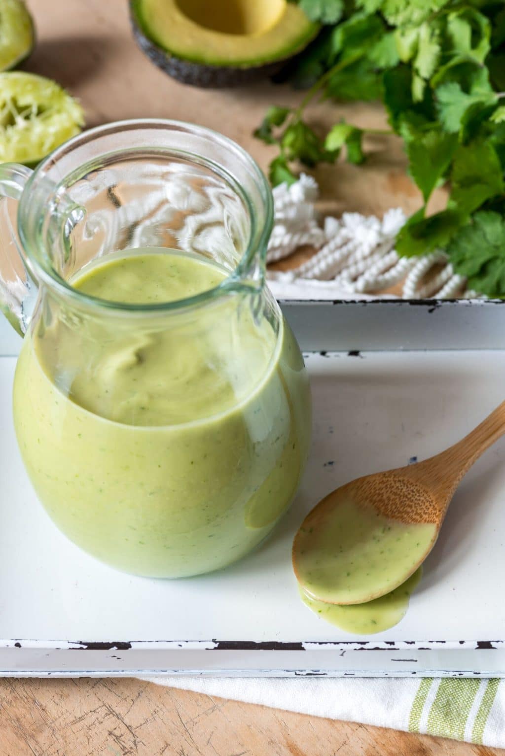 a glass pitcher filled with creamy avocado sauce on a white and green towel with a wooden spoon on the side with sauce dripping off of it and avocados, parsley and a half of a squeezed lime in the background