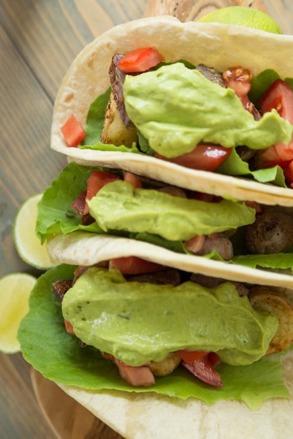 Three potato tacos with tomato and a large dollop of avocado lime sauce on a wood board with lime wedges next to them