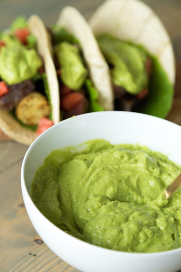 Creamy Avocado Sauce in a white bowl with three potato tacos topped with the sauce in the background