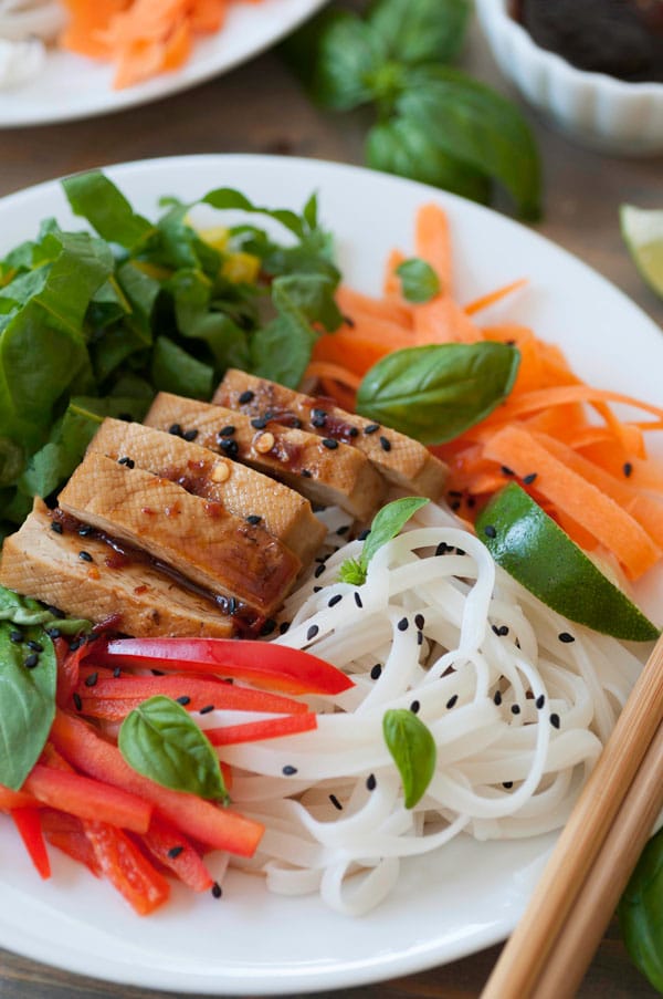 Chilled rice noodles, bell pepper slices, carrots, smoked tofu, chard and basil on a white plate, sprinkled with black sesame seeds