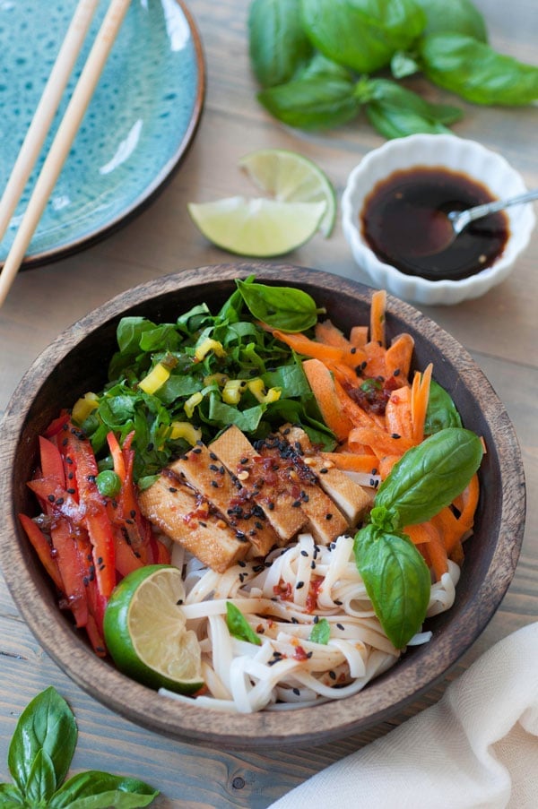 A wood bowl of chilled sesame noodle salad with a small white bowl of dressing, plates, chopsticks, and basil leaves and a lime wedge in the background.