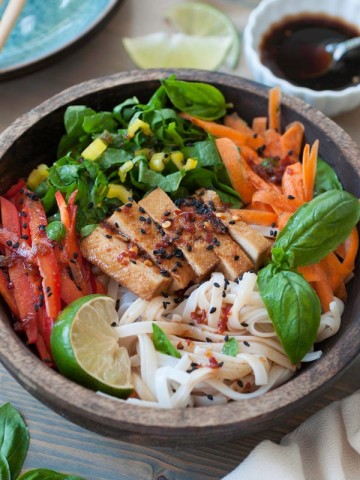 A wood bowl with cold sesame noodles and slivered bell peppers, carrots and yellow chard with basil and a lime wedge and a small white bowl of sesame dressing in the background with stacked blue plates and chopsticks