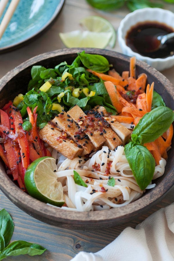 A wood bowl with cold sesame noodles and slivered bell peppers, carrots and yellow chard with basil and a lime wedge and a small white bowl of sesame dressing in the background with stacked blue plates and chopsticks