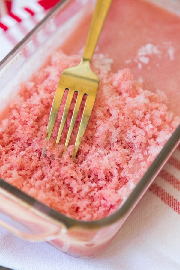 Frozen lemon watermelon granita in a glass bread pan being scraped with a gold fork