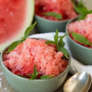 Three green bowls of lemon watermelon granita with a stack of silver spoons and a watermelon wedge on a wood table
