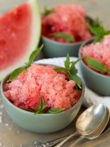 Three green bowls of lemon watermelon granita with a stack of silver spoons and a watermelon wedge on a wood table