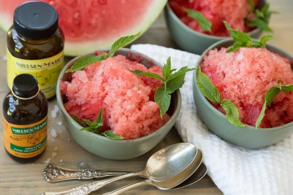 Three green bowls filled with lemon watermelon granita with silver spoons a white cloth, a wedge of watermelon and a bottle of Nielsen-Massey Pure Lemon Paste and Pure Organic Orange Extract