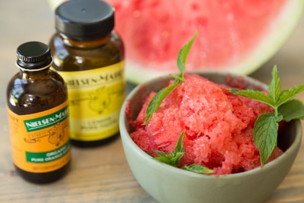 A green bowl of lemon watermelon granita with mint leaves and a bottle of Nielsen-Massey Pure Lemon Paste and Pure Organic Orange Extract and a watermelon wedge