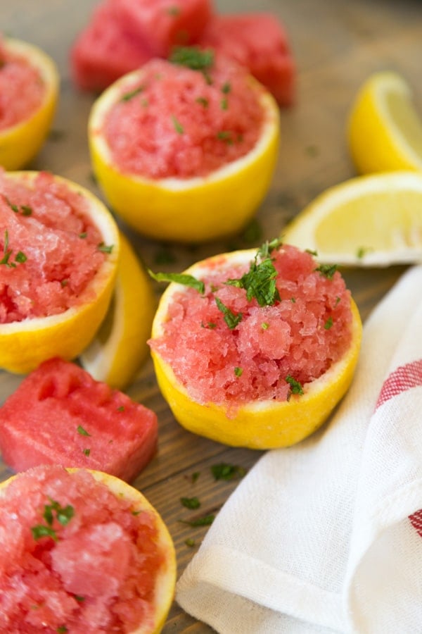 Four lemon cups filled with lemon watermelon granita with mint leaves on top and pieces of watermelon and lemon wedges scattered on a wood board