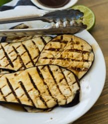 Grilled eggplant with lime zest on a white plate with tongs on a wood board