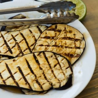 Grilled eggplant with lime zest on a white plate with tongs on a wood board