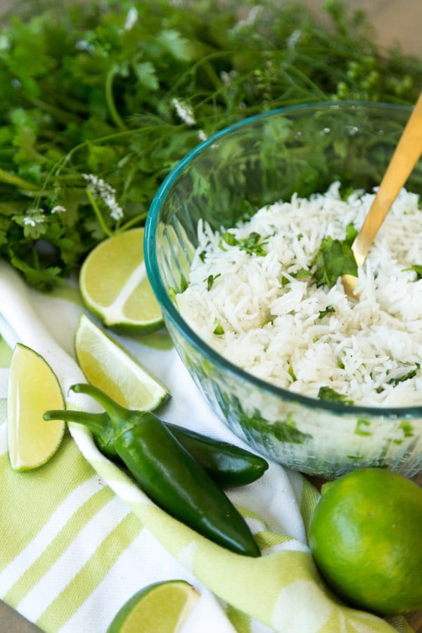 A glass bowl filled with cilantro lime rice on a green and white towel with a gold spoon in the bowl with jalapeno peppers, limes and a bunch of cilantro around it. 