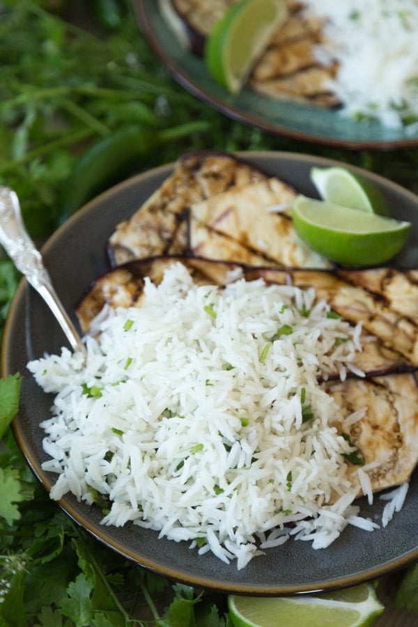 A pile of cilantro lime rice with grilled eggplant on a dark plate with a silver fork and lime wedges