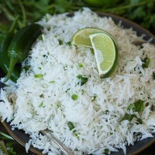 A pile of cilantro lime Mexican Rice on a dark plate with a lime wedge and cilantro on top and a silver fork next to the plate