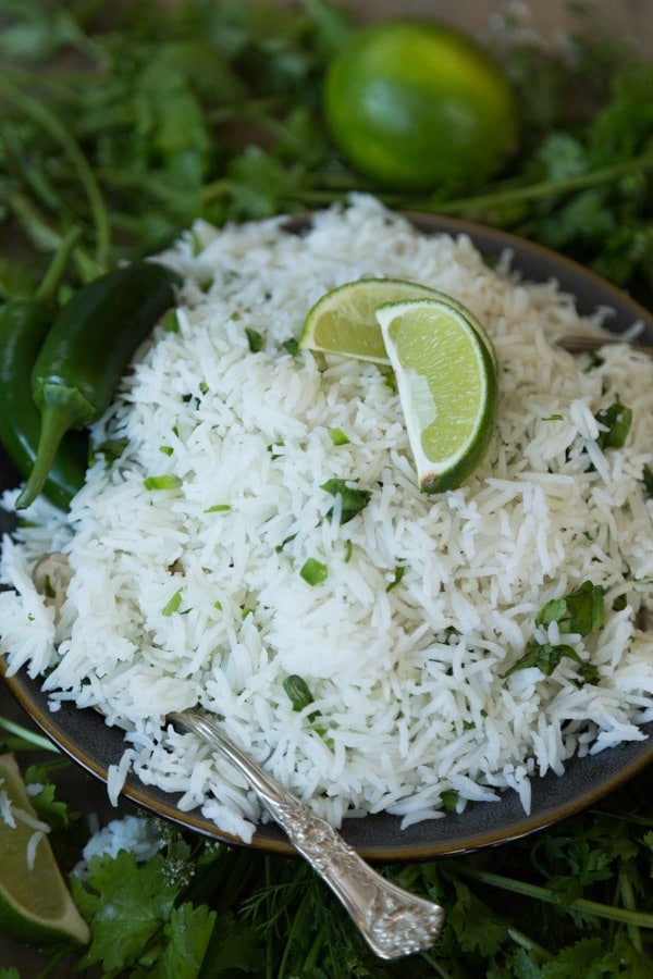 A pile of cilantro lime Mexican Rice on a dark plate with a lime wedge and cilantro on top and a silver fork next to the plate