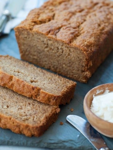 A loaf of vegan banana bread with two slices cut off the end on a slate board with a wood bowl of vegan butter and a butter knife next to the bread