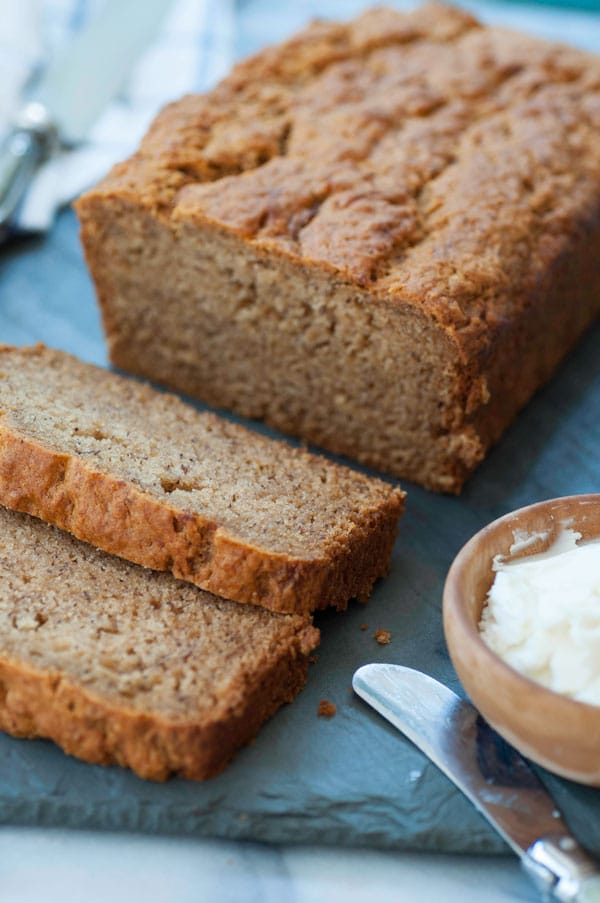 A loaf of vegan banana bread with two slices cut off the end on a slate board with a wood bowl of vegan butter and a butter knife next to the bread