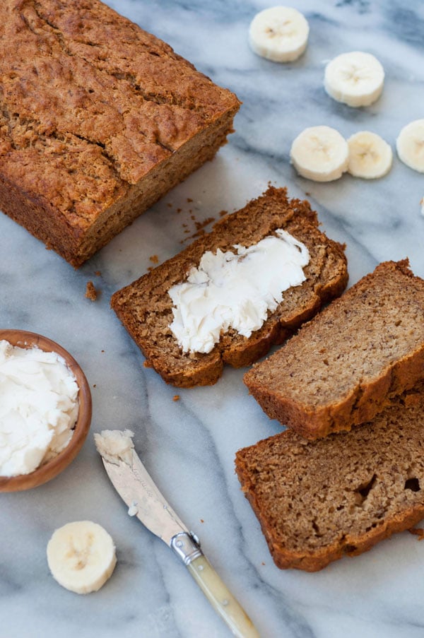 A loaf of vegan banana bread on a marble board with three slices scattered on the board, one of them with a smear of vegan butter and a wood bowl of butter and a butter knife and banana slices scattered around the bread