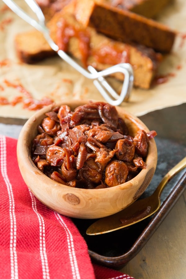 Cherry confit in a wood bowl with a red napkin, gold spoon, and vegan ribs in the background