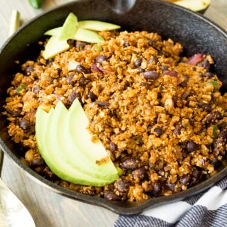 Vegan taco filling in an iron skillet with avocado slices on top and halved avocados and a jalapeno next to the pan