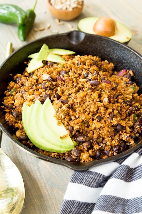 Vegan taco filling in an iron skillet with avocado slices on top and halved avocados and a jalapeno next to the pan