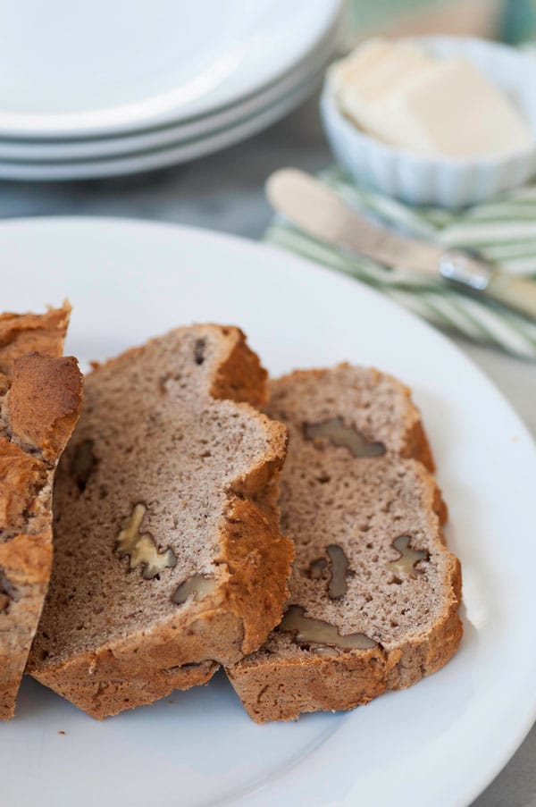 A plate with a loaf of sliced banana bread and three white plates in the background with a dish of butter and a butter knife on a green striped napkin