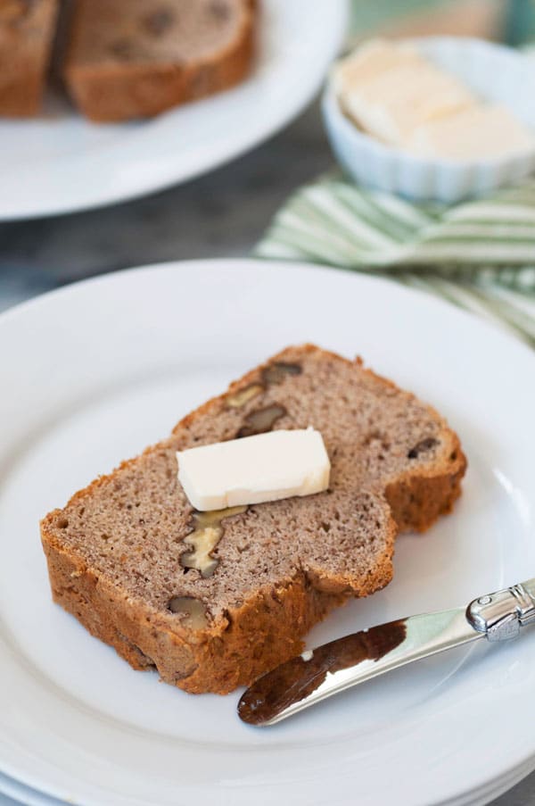 A slice of banana bread with a pat of butter on top on a white plate with a butter knife on the side of the plate and a plate of bread in the background with a green striped towel and a white dish of butter