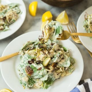 Brussels sprouts salad with mixed greens covered in Caesar dressing on a white plate with a gold fork full of salad and two white plates with salad on the sides and lemon wedges