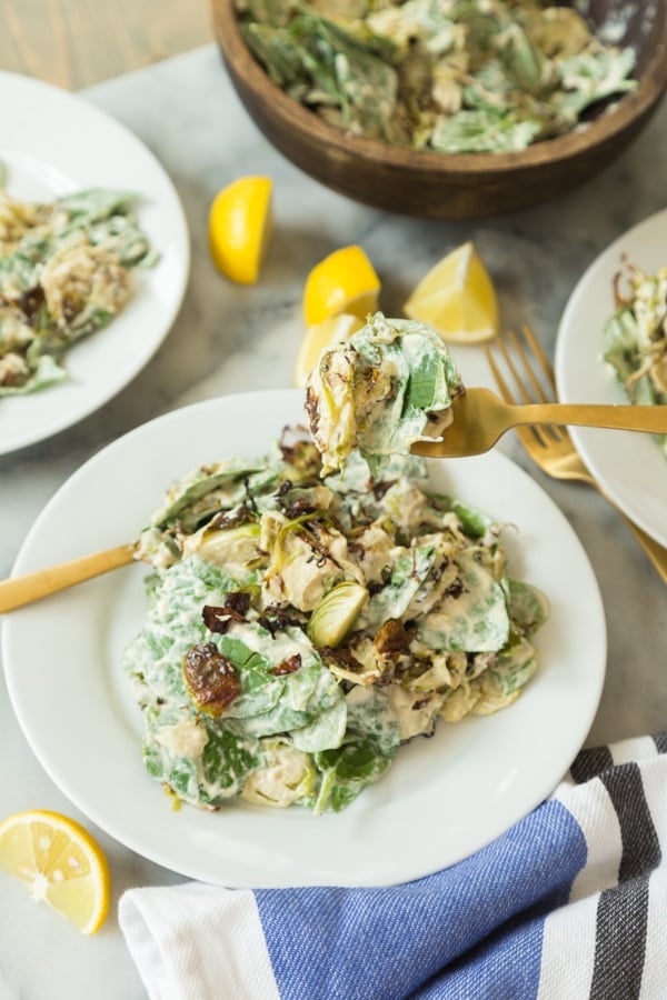 Brussels sprouts salad with mixed greens covered in Caesar dressing on a white plate with a gold fork full of salad and two white plates with salad on the sides and lemon wedges