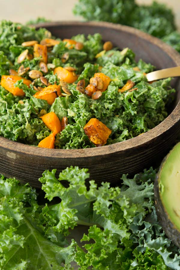 Kale and roasted squash salad in a wood bowl with a gold fork in the bowl and kale leaves and a sliver of avocado next to the bowl
