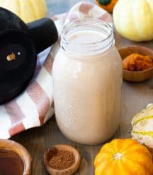 A mason jar filled with pumpkin spice cashew milk with brown wood bowls of spices and maple syrup next to it, and pumpkins surrounding the jar