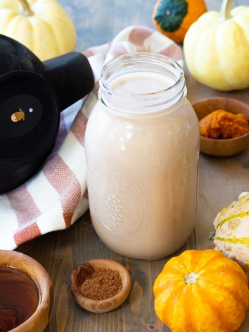 A mason jar filled with pumpkin spice cashew milk with brown wood bowls of spices and maple syrup next to it, and pumpkins surrounding the jar