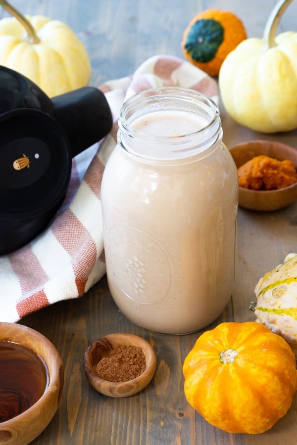 A mason jar filled with pumpkin spice cashew milk with brown wood bowls of spices and maple syrup next to it, and pumpkins surrounding the jar