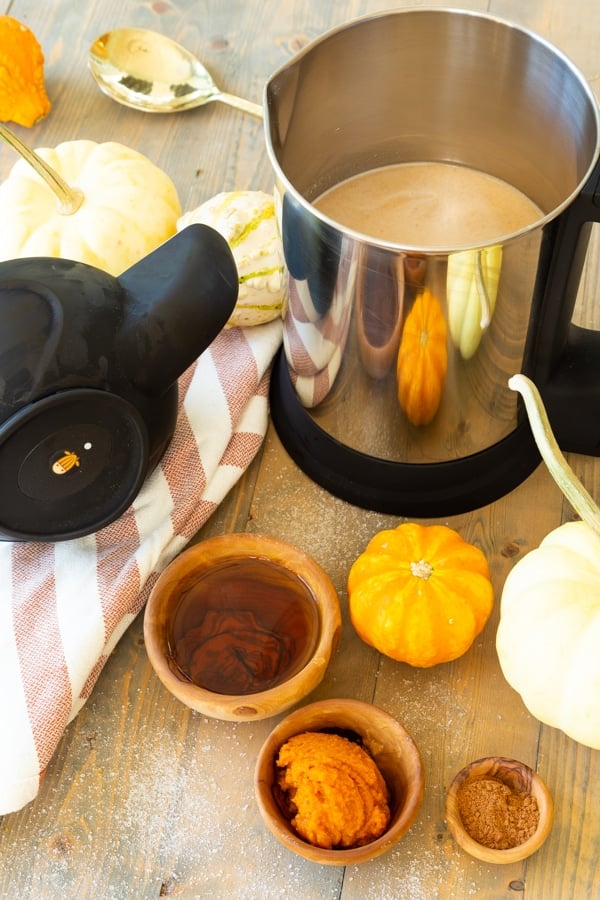 The Almond Cow milk maker on a wood board with the ingredients (pumpkin puree, pumpkin spice, maple syrup) and pumpkins around it. 