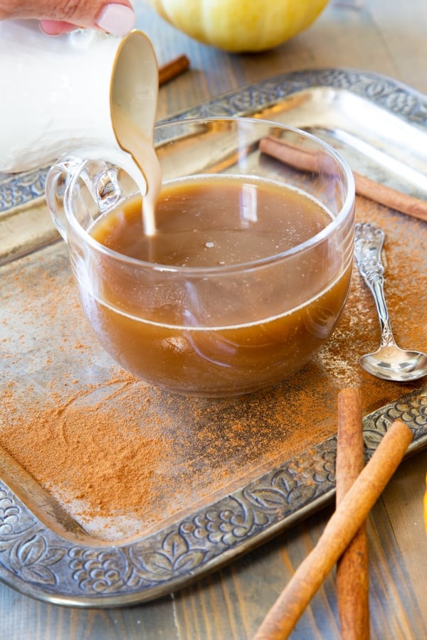 A big glass mug of coffee with pumpkin spice cashew milk being poured into the cup on a silver tray with cinnamon sprinkled around and cinnamon sticks next to it with a silver spoon.