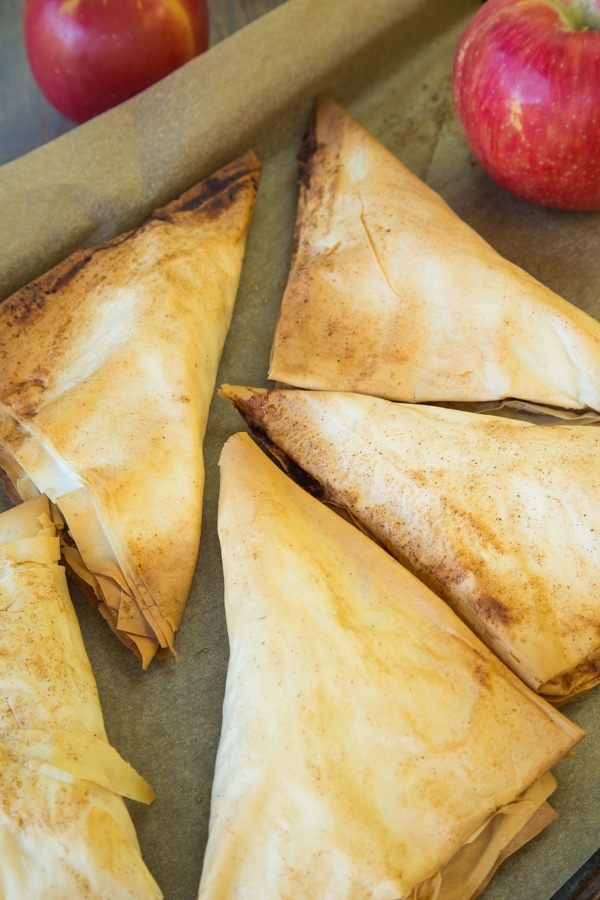 Several brown and crispy apple turnovers on a baking sheet with parchment paper and red apples next to them