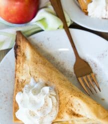 A vegan apple turnover with a dollop of whipped cream on top on a white plate with a gold fork on it and a red apple next to the plate