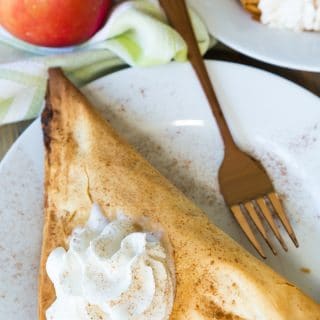 A vegan apple turnover with a dollop of whipped cream on top on a white plate with a gold fork on it and a red apple next to the plate