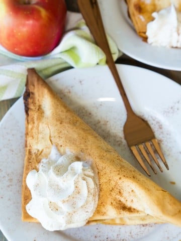 A vegan apple turnover with a dollop of whipped cream on top on a white plate with a gold fork on it and a red apple next to the plate