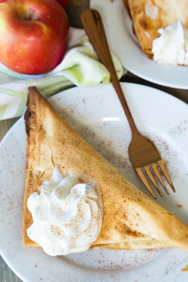 A vegan apple turnover with a dollop of whipped cream and cinnamon sprinkled on top on a white plate with a gold fork on it and a red apple next to the plate