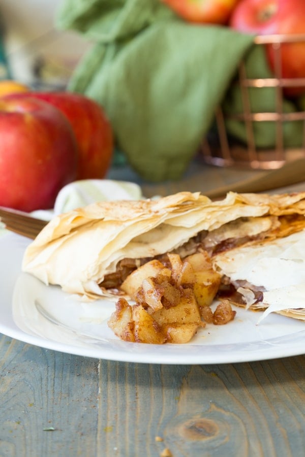 An apple turnover cut in half with apple filling spilling from the center on a white plate with a copper basket of apple and a green towel in the background