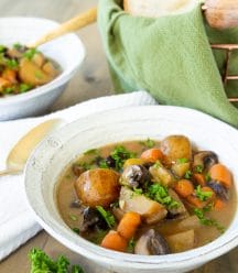 Two bowls of vegan beef stew in white bowls with sprigs of parsley and a copper wire bread basket with a green towel and bread in the background