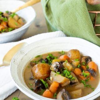 Two bowls of vegan beef stew in white bowls with sprigs of parsley and a copper wire bread basket with a green towel and bread in the background