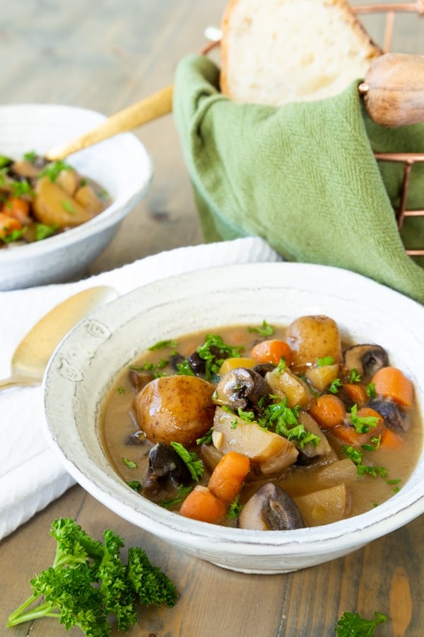 Two bowls of vegan beef stew in white bowls with sprigs of parsley and a copper wire bread basket with a green towel and bread in the background