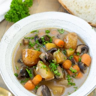 A bowl of vegan beef stew in a white bowl with a slice of bread and a gold spoon and a sprig of parsley on the side