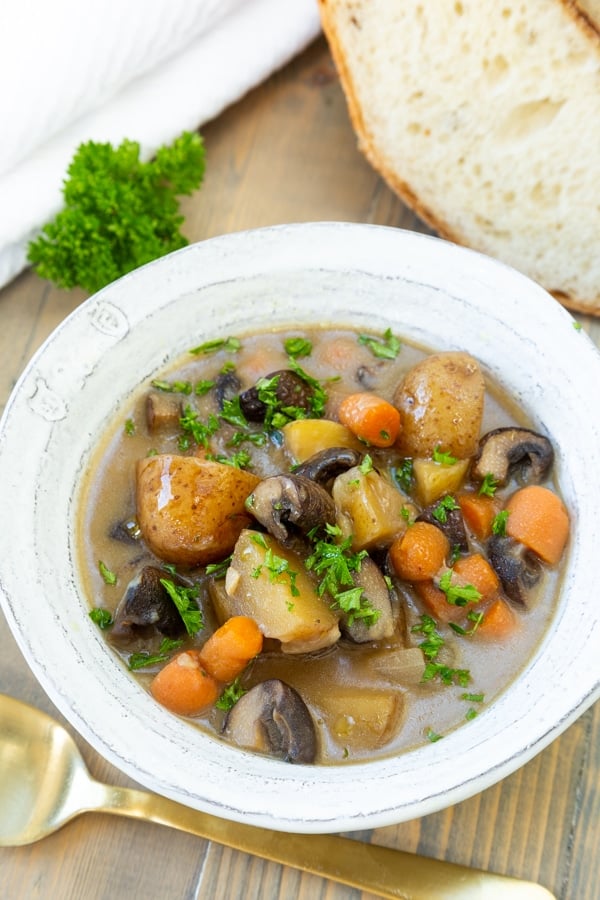 A bowl of vegan beef stew in a white bowl with a slice of bread and a gold spoon and a sprig of parsley on the side