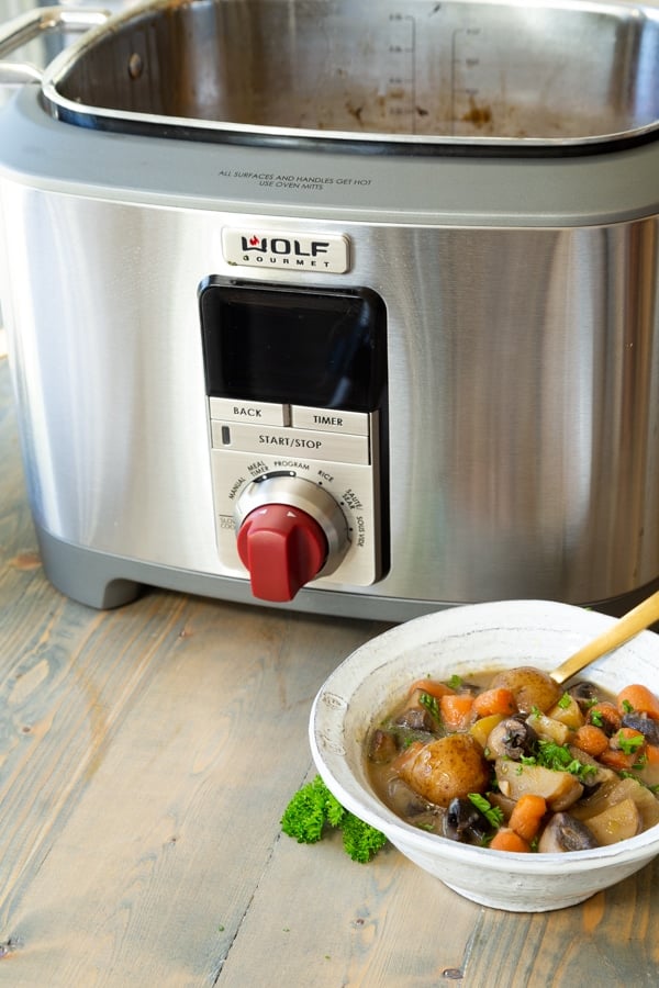 A bowl of vegan beef stew in a white bowl with a gold spoon in the bowl in front of a Wolf Gourmet Multi-Function Cooker simmering a pot of the stew