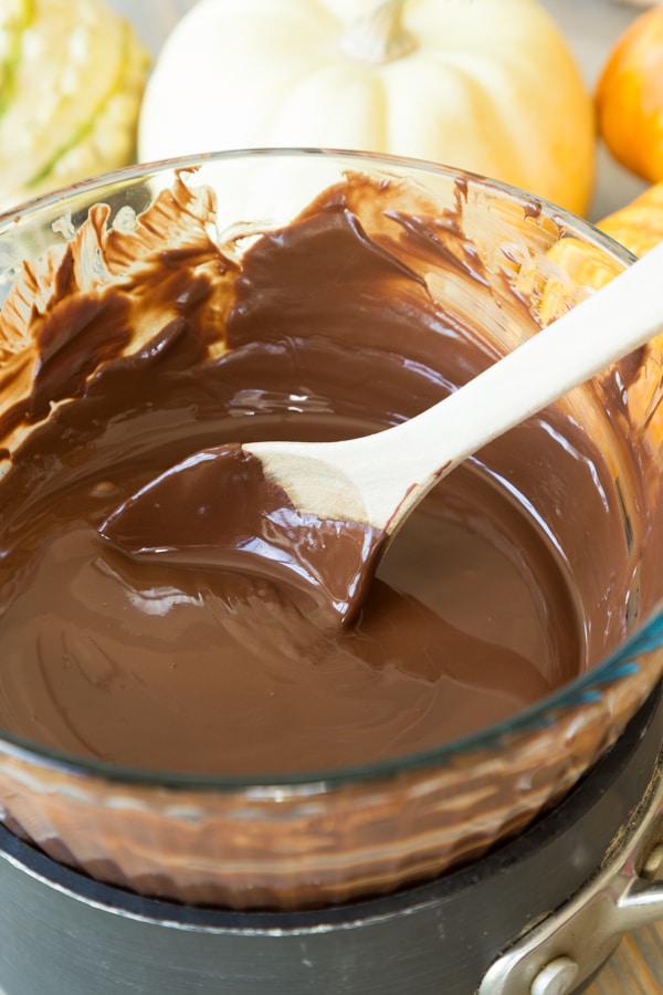 A glass bowl full of melted chocolate over a sauce pan with a wooden spoon stirring the chocolate, with white and orange pumpkins in the background