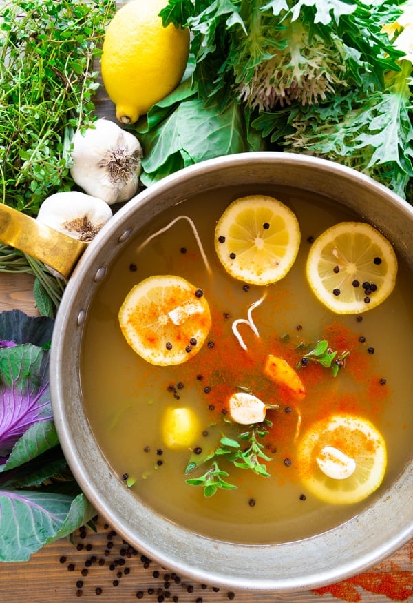 A pot of vegetable broth with lemons, garlic, turmeric, fresh herbs and peppercorns floating on top