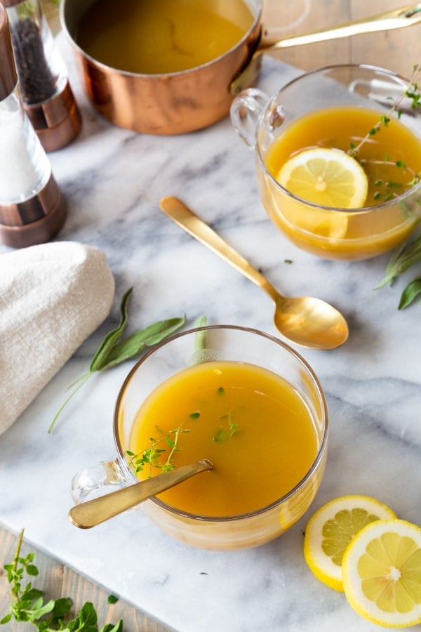 Two glass bowls of vegetable broth with gold spoons and the pot of broth and ingredients next to the bowls on a marble board